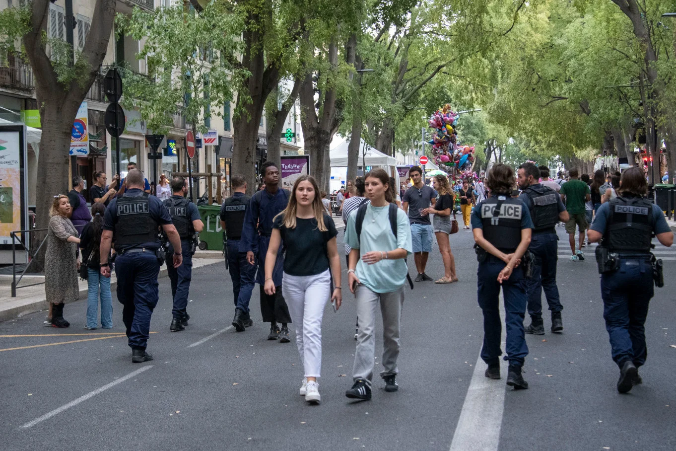 Plusieurs policiers en uniforme patrouillent dans une rue piétonne animée sous de grands arbres.