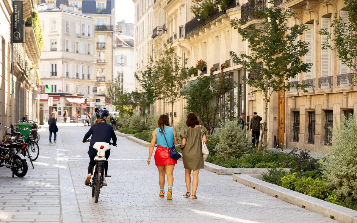 Une cycliste et des piétons parcourent une rue parisienne pavée agrémentée de végétalisation urbaine.