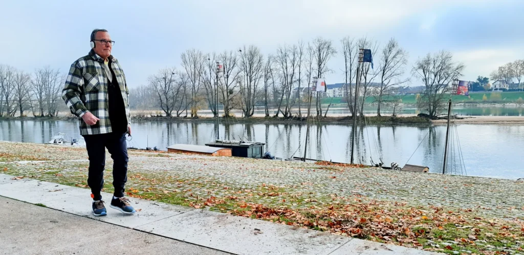 Un homme âgé en veste à carreaux marche sur un quai pavé bordant une rivière calme.