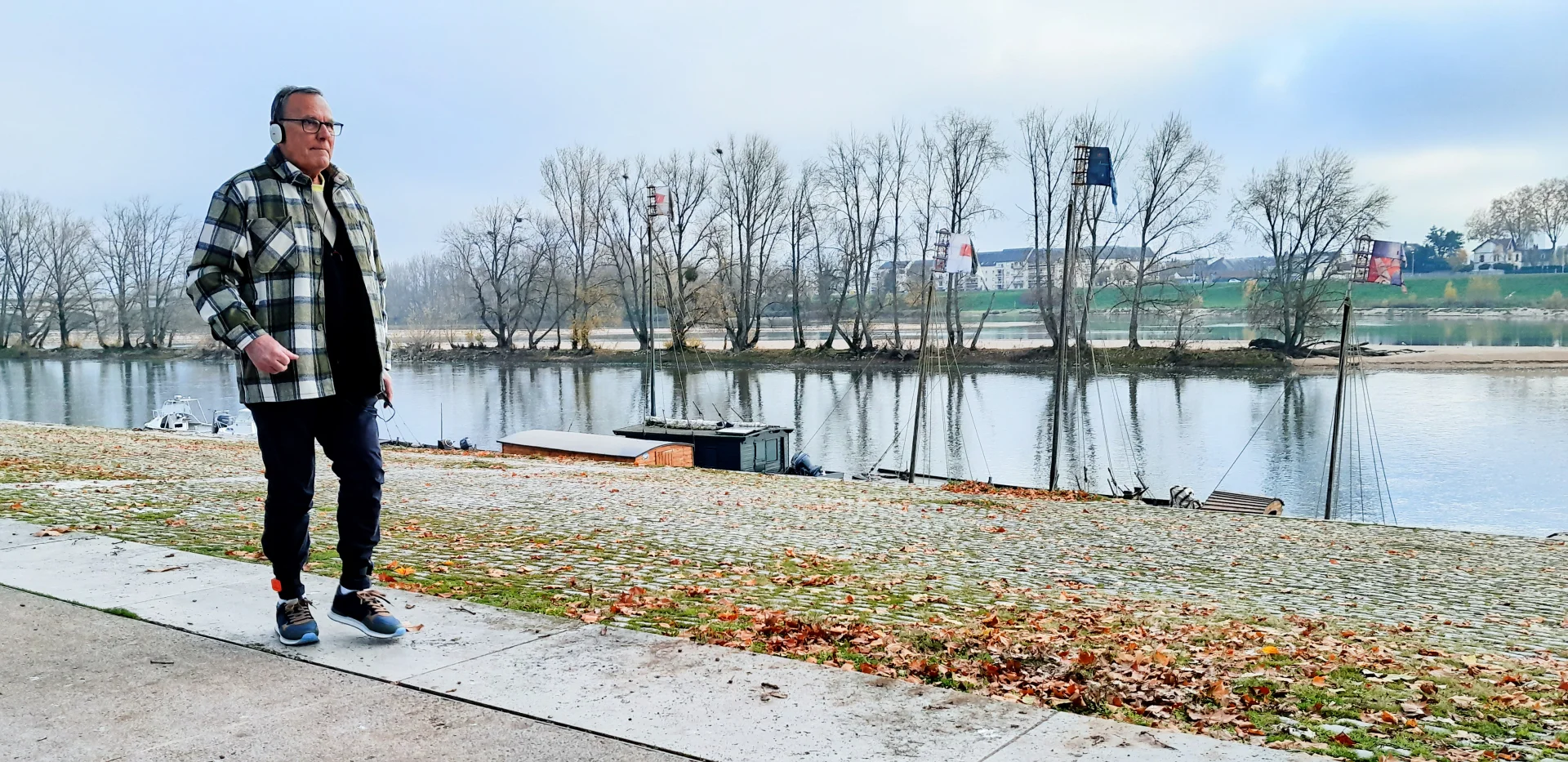 Un homme âgé en veste à carreaux marche sur un quai pavé bordant une rivière calme.
