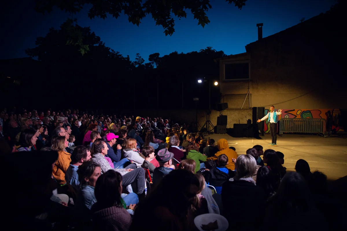Un artiste en gilet bleu se produit sous un projecteur devant une foule attentive nocturne.
