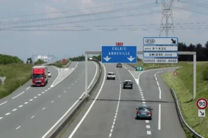 Voitures et camion rouge circulant sur une autoroute sous des panneaux directionnels vers Calais.