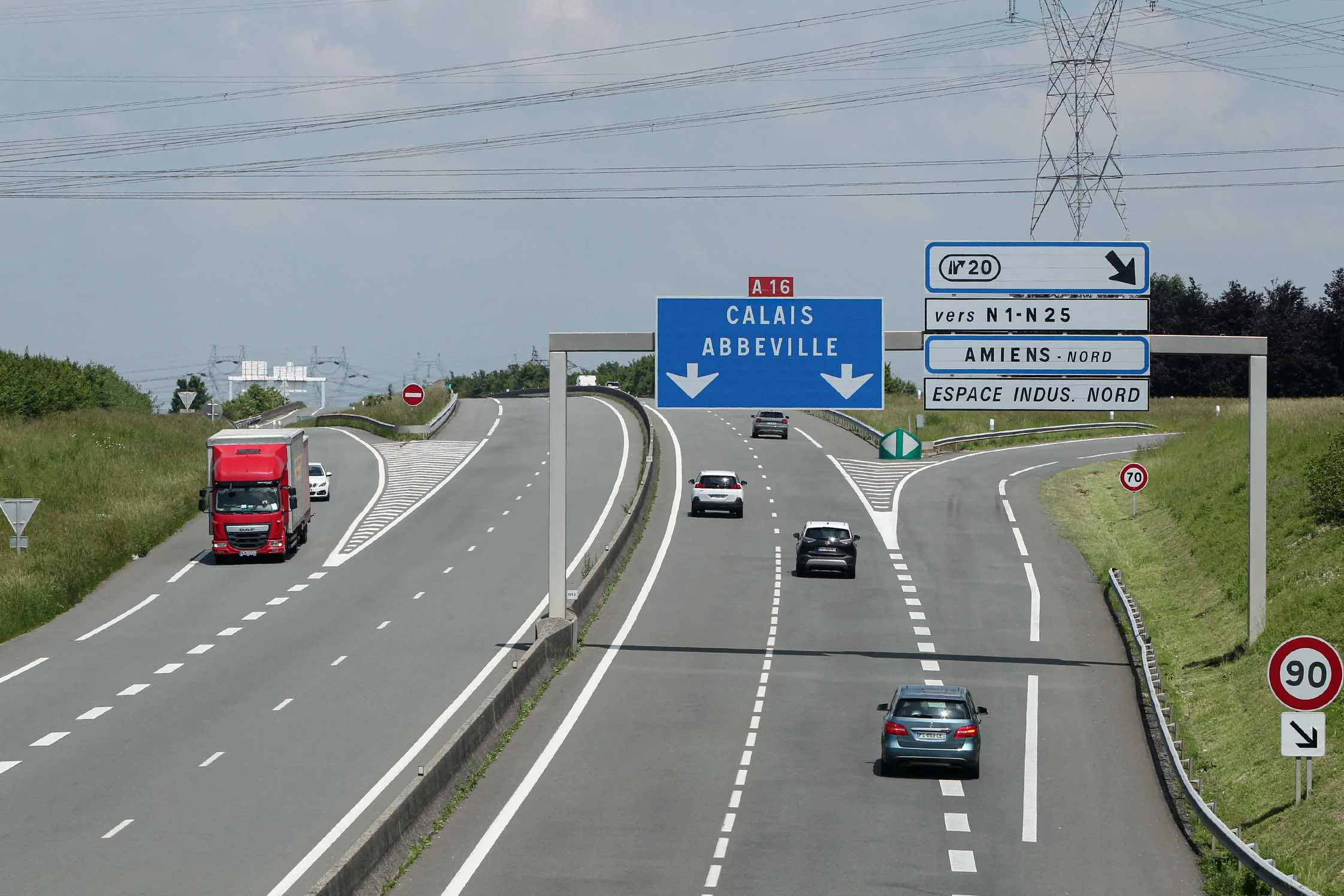 Voitures et camion rouge circulant sur une autoroute sous des panneaux directionnels vers Calais.