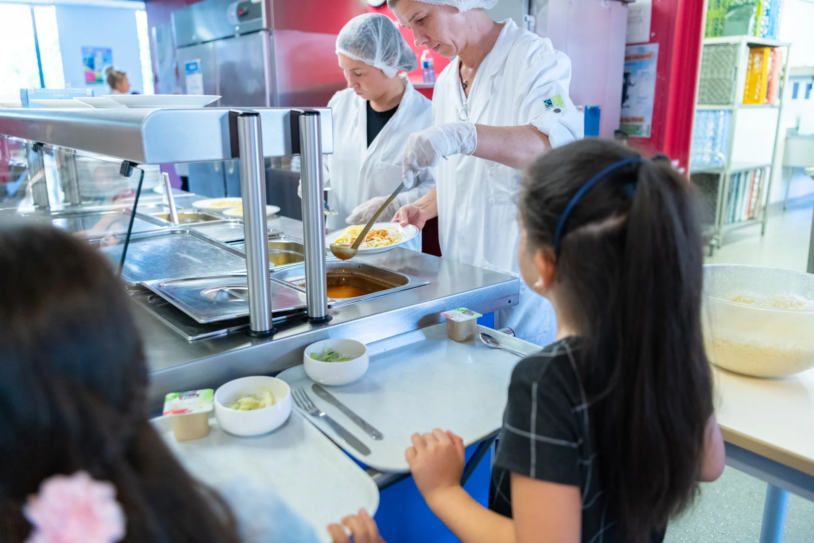 Une employée de cantine en blouse blanche sert des pâtes à une écolière dans un réfectoire.