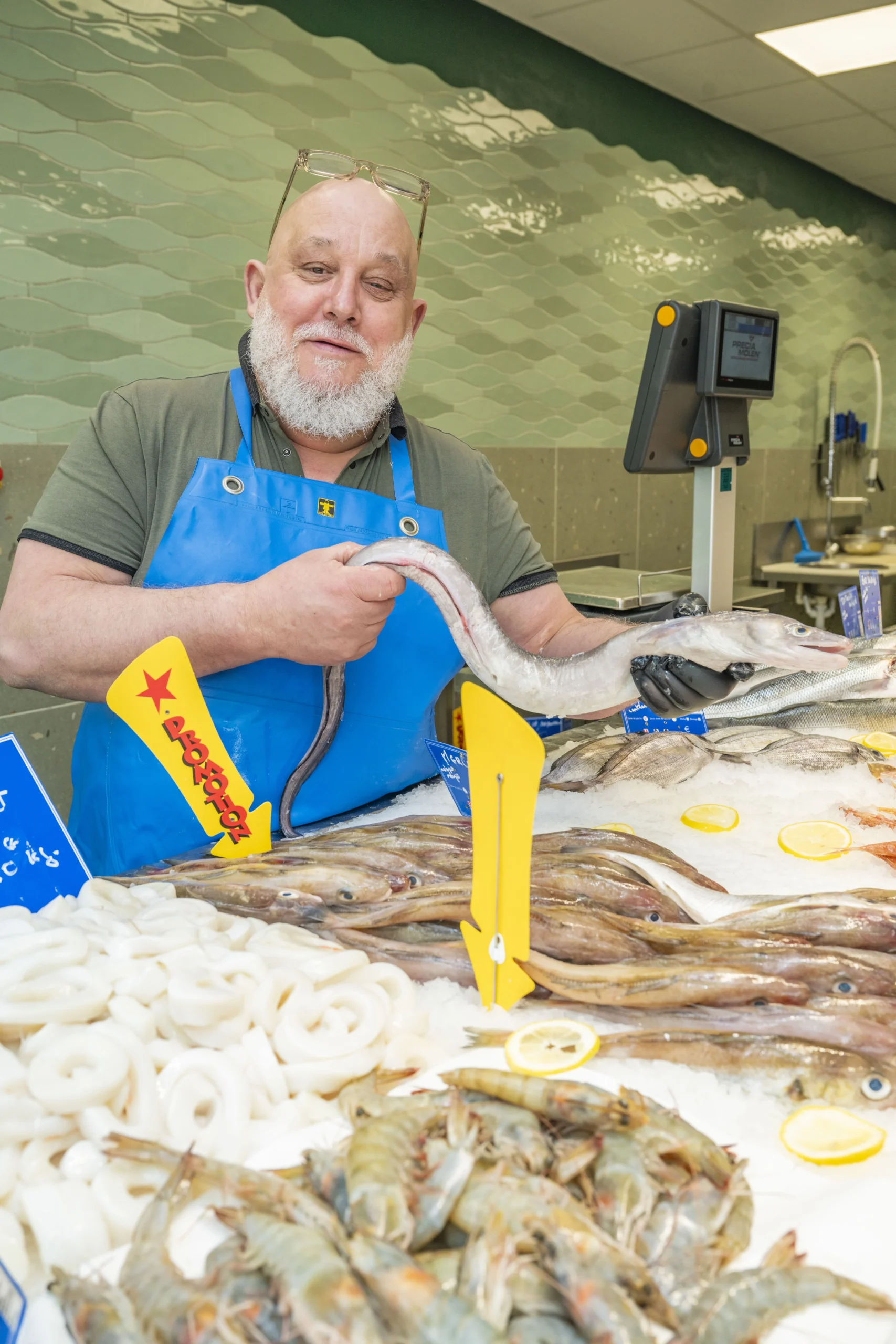 Un poissonnier barbu présente un long poisson argenté au-dessus d'un étalage de fruits de mer.