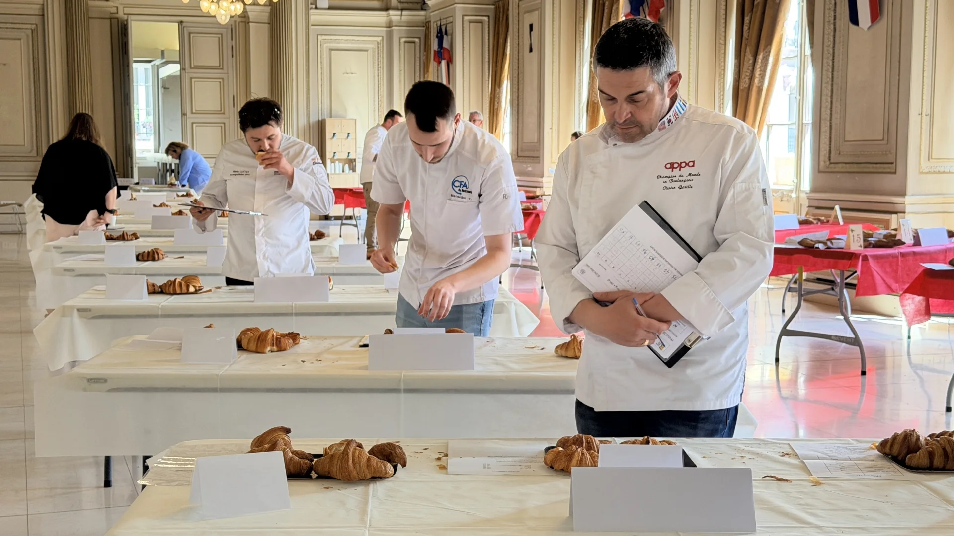 Des boulangers en vestes blanches évaluent des croissants disposés sur des tables lors d'un concours.