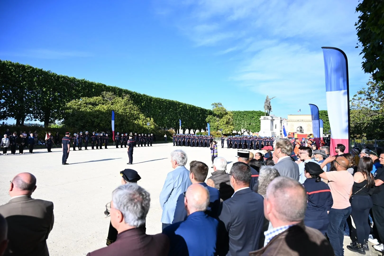 Pompiers en uniforme alignés sur une place publique devant une foule lors d'une cérémonie officielle.