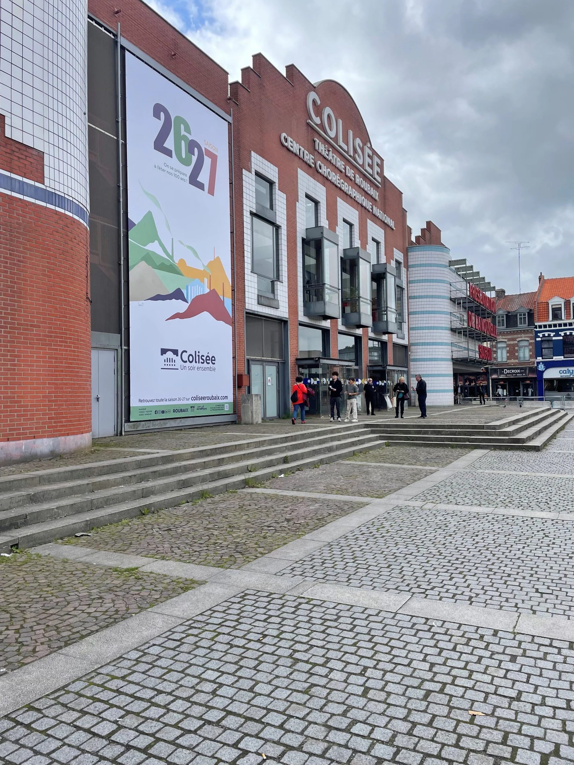 Façade en briques du théâtre Le Colisée à Roubaix avec une affiche géante devant l'entrée.