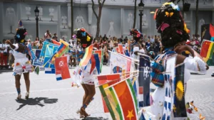 Plusieurs danseuses en costume blanc défilent dans la rue, portant de nombreux drapeaux de différents pays fixés à leurs vêtements, symbolisant la diversité culturelle.