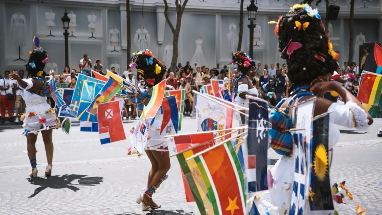 Plusieurs danseuses en costume blanc défilent dans la rue, portant de nombreux drapeaux de différents pays fixés à leurs vêtements, symbolisant la diversité culturelle.