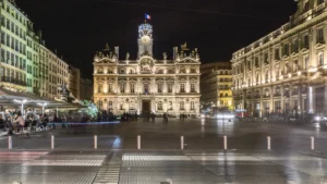 Une place publique française illuminée de nuit avec un bâtiment historique majestueux en arrière-plan.