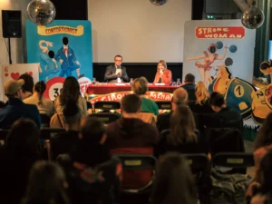 Deux personnes s'expriment à une table devant un public, entourées de décors de cirque vintage.