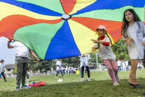 Enfants jouant sous un grand parachute multicolore sur un terrain de sport ensoleillé.