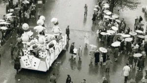 Char de défilé orné de cartes à jouer et personnages géants devant une foule sous parapluies.