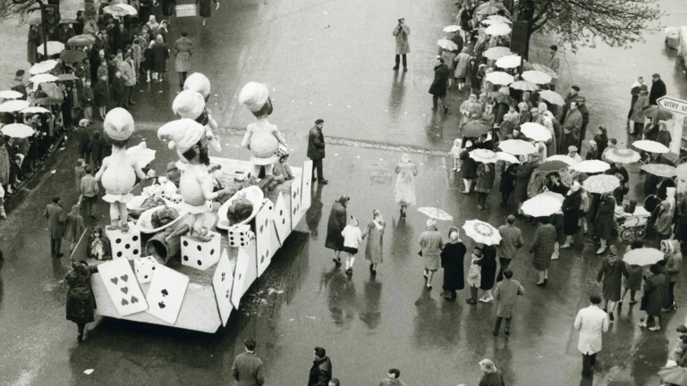 Char de défilé orné de cartes à jouer et personnages géants devant une foule sous parapluies.