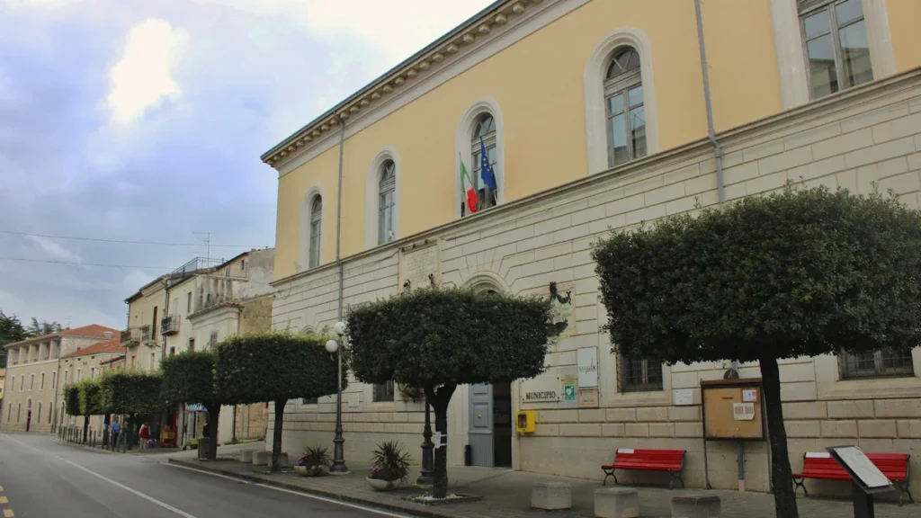 Façade d'un bâtiment municipal aux murs jaunes avec des arbres décoratifs et des bancs rouges.