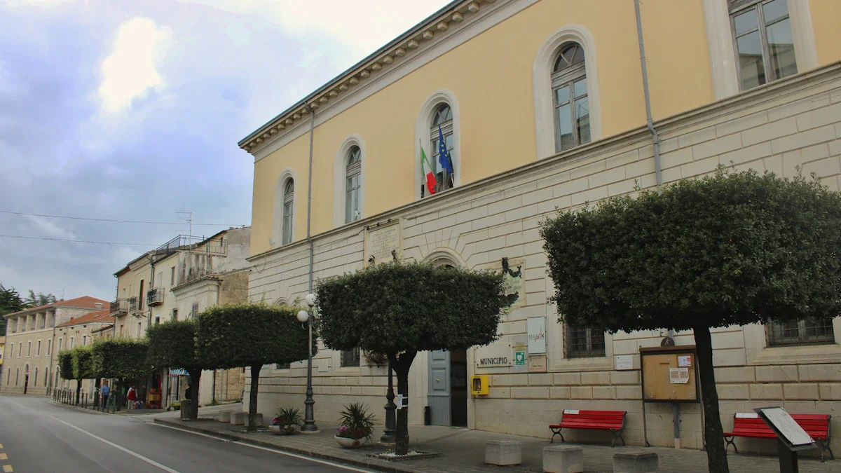 Façade d'un bâtiment municipal aux murs jaunes avec des arbres décoratifs et des bancs rouges.
