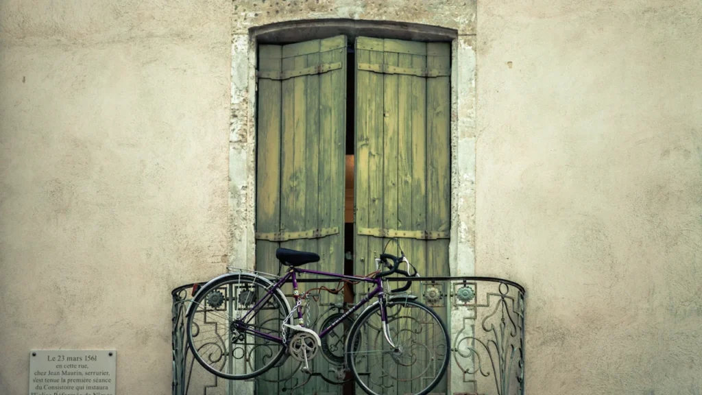 Un vélo accroché à un balcon en fer forgé devant des volets en bois.