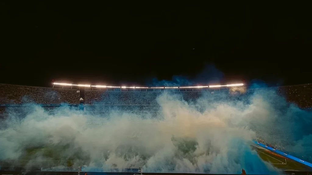 Vue nocturne d'un stade de football rempli de fumigènes bleus et d'ambiance festive.