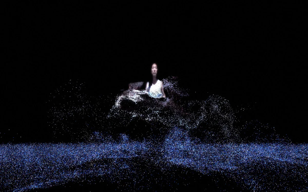 Une silhouette féminine émerge d'un nuage de particules lumineuses bleues sur fond noir.