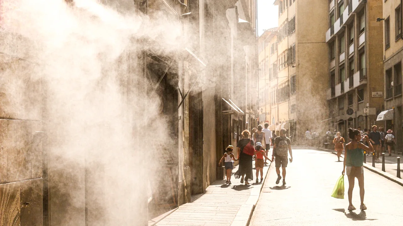 Des passants marchent dans une rue ensoleillée sous une brume rafraîchissante pendant la canicule.