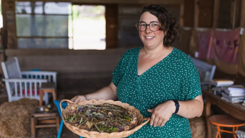 Une femme souriante aux cheveux courts tenant un plateau en osier rempli d'herbes séchées.