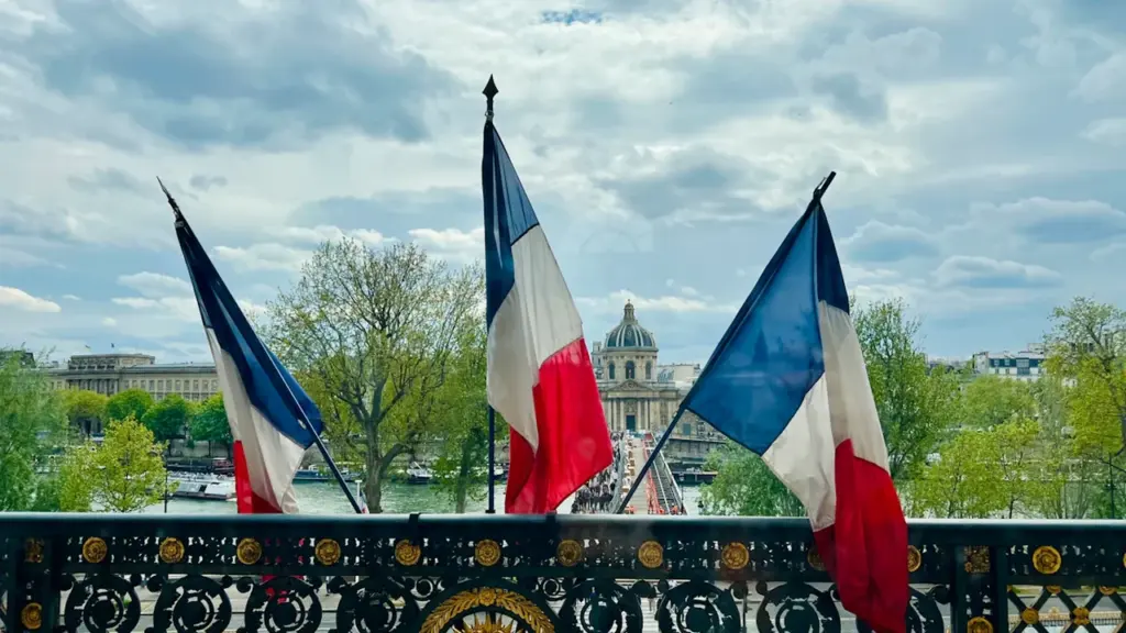 Trois drapeaux français flottant au-dessus d'une balustrade avec une architecture parisienne en arrière-plan.
