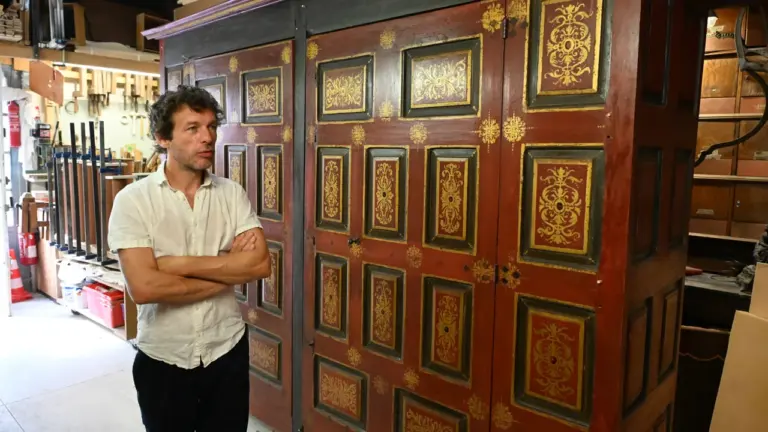 Un ébéniste se tient devant une grande armoire ancienne en bois rouge ornée de dorures.