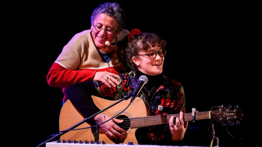Une femme mature pose sa main sur l'épaule d'une jeune guitariste sur scène sombre.