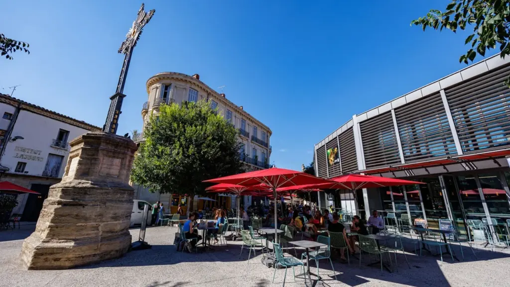 Une place ensoleillée avec une grande croix en pierre, des parasols rouges et des passants.