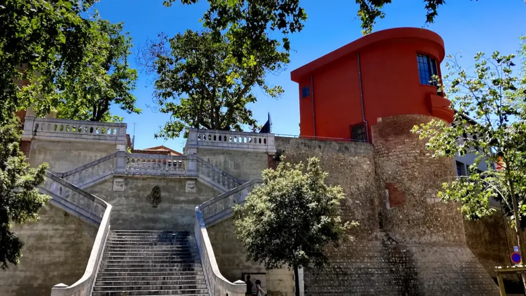 Un escalier monumental en pierre menant vers un bâtiment historique rouge à Perpignan.