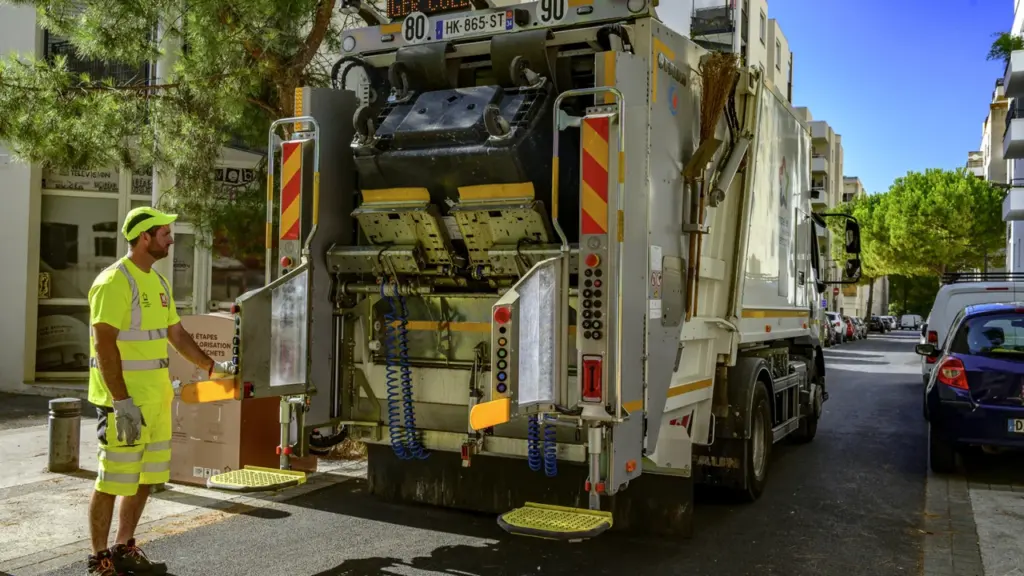 Un agent de collecte des déchets en tenue fluorescente près de son camion dans la rue.