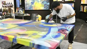 Un artiste barbu en t-shirt blanc peint une grande toile colorée dans une galerie d'art.