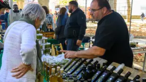 Un commerçant conseille une cliente sur des bouteilles d'huile d'olive sur un marché extérieur.